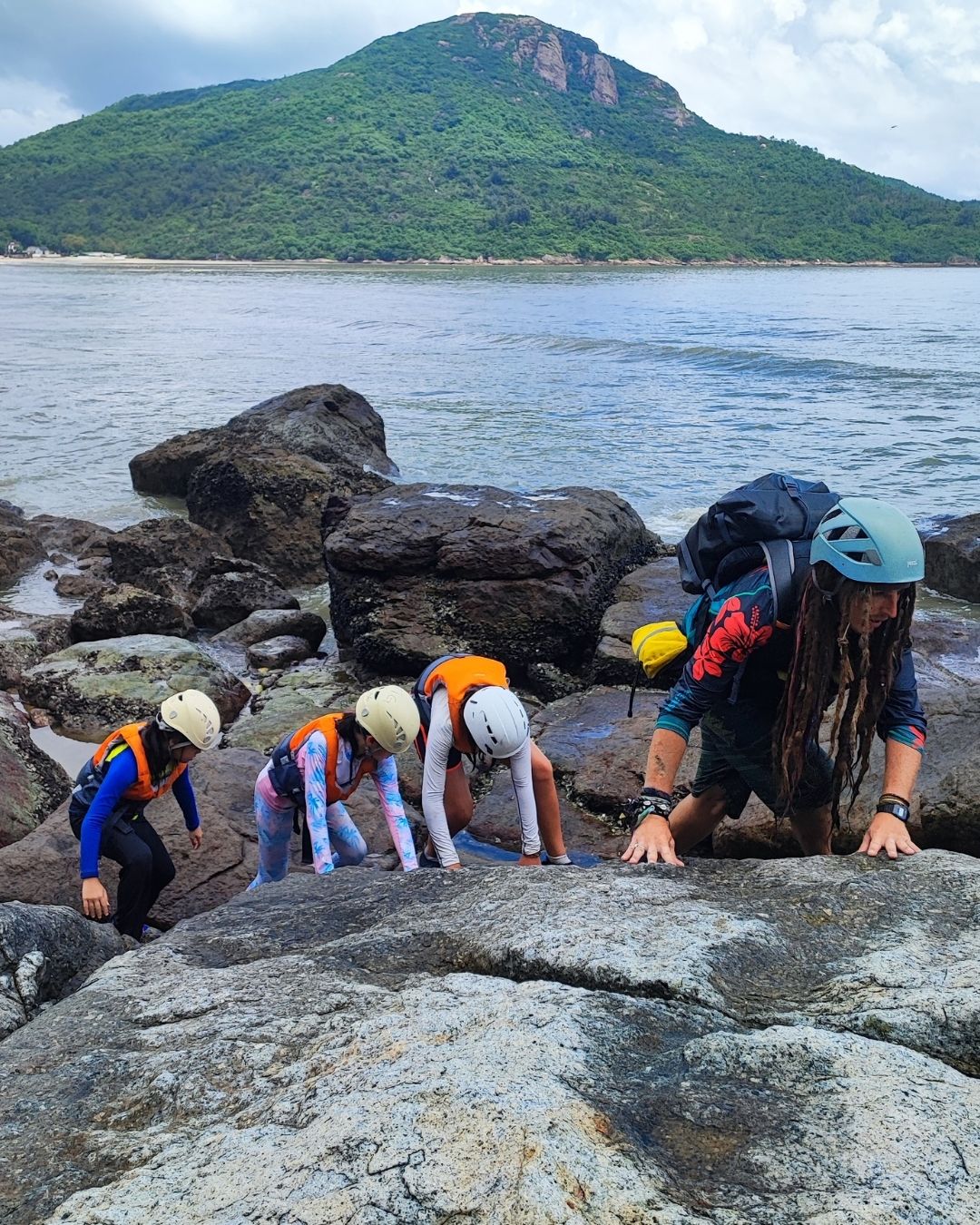 Group of people exploring rocky terrain by a body of water with a mountain in the background.